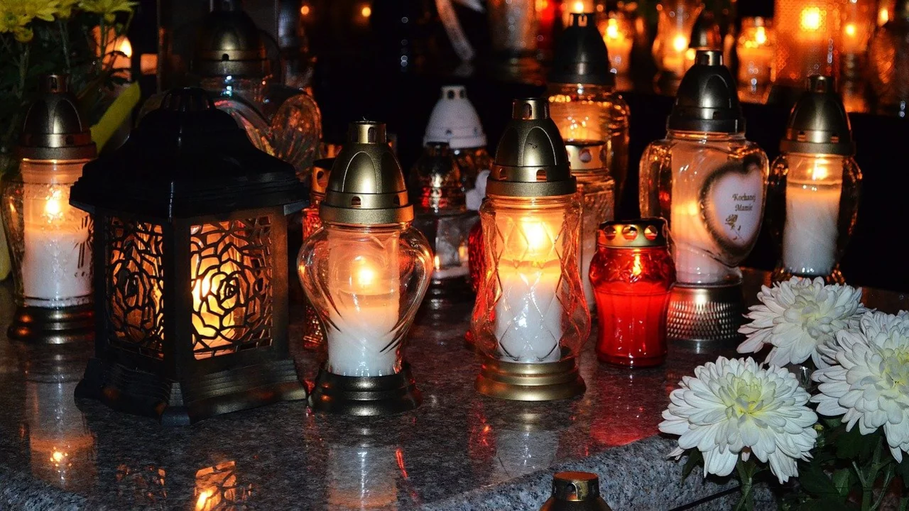 Eine Ansammlung von verschiedenen Grablichtern steht auf einem Steinfeld. Die Lichter leuchten in warmen Farben, umgeben von Blumen. Die Szene vermittelt eine friedliche und respektvolle Atmosphäre, die an einen Gedenkort erinnert.