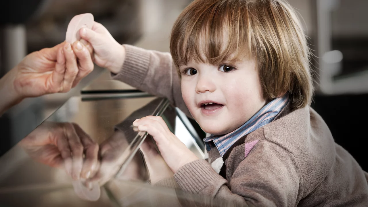 Ein kleines Kind mit blonden Haaren lächelt und greift nach einem Gegenstand, der von einer Hand gereicht wird. Es sitzt an einem Tisch mit reflektierender Oberfläche, was den Moment freundlich und einladend wirken lässt.