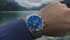 Ein Handgelenk hält eine Uhr mit einem blauen Zifferblatt und silbernem Armband. Im Hintergrund ist eine ruhige Gewässerlandschaft mit Bergen und bewölktem Himmel zu sehen. Die Uhr ist klar im Fokus und vermittelt ein Gefühl von Zeit und Abenteuer.