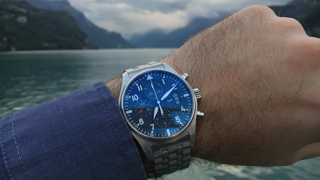 Ein Handgelenk hält eine Uhr mit einem blauen Zifferblatt und silbernem Armband. Im Hintergrund ist eine ruhige Gewässerlandschaft mit Bergen und bewölktem Himmel zu sehen. Die Uhr ist klar im Fokus und vermittelt ein Gefühl von Zeit und Abenteuer.