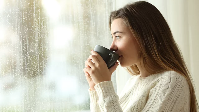 Eine junge Frau sitzt in einem hellen Raum und hält eine Tasse Kaffee in der Hand. Sie blickt nachdenklich aus dem Fenster, während es draußen regnet. Ihr Ausdruck vermittelt Entspannung und ein Gefühl von Gemütlichkeit.