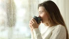 Eine junge Frau sitzt in einem hellen Raum und hält eine Tasse Kaffee in der Hand. Sie blickt nachdenklich aus dem Fenster, während es draußen regnet. Ihr Ausdruck vermittelt Entspannung und ein Gefühl von Gemütlichkeit.