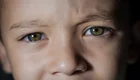 Nahaufnahme eines Kindes mit leuchtend grünen Augen und einem nachdenklichen Gesichtsausdruck. Die Haut des Kindes ist leicht gebräunt. Der Hintergrund ist dunkel, wodurch die Augen besonders hervorstechen.