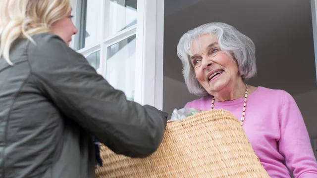 Eine lächelnde ältere Frau steht an einem Fenster und empfängt eine jüngere Frau, die einen großen Korb hält. Die ältere Dame trägt einen pinken Pullover und scheint erfreut über den Besuch zu sein.
