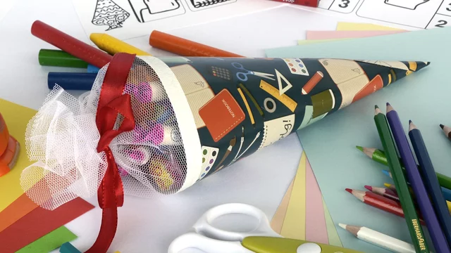 Das Bild zeigt eine traditionelle Schultüte in Form einer gefüllten, bunten Papiertüte. Um die Tüte herum sind Bastelmaterialien wie Buntstifte, Papier und eine Locher zu sehen. Die Schultüte symbolisiert den Beginn des neuen Schuljahres für Kinder.