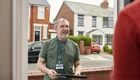 Ein lächelnder Mann steht an einer Haustür, hält ein Tablett in der Hand und trägt ein Ausweis. Im Hintergrund sind Wohnhäuser und Autos zu sehen. Der Mann scheint eine Lieferung oder einen Besuch zu machen.