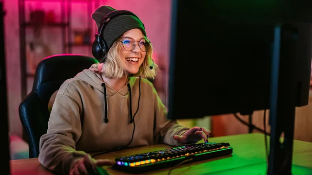 Eine lächelnde junge Frau sitzt am Computer und spielt Videospiele. Sie trägt einen grauen Hoodie, eine Mütze und ein Headset. Vor ihr befindet sich eine Tastatur und der Bildschirm zeigt vermutlich ein Spiel. Die Beleuchtung im Hintergrund ist farbenfroh.