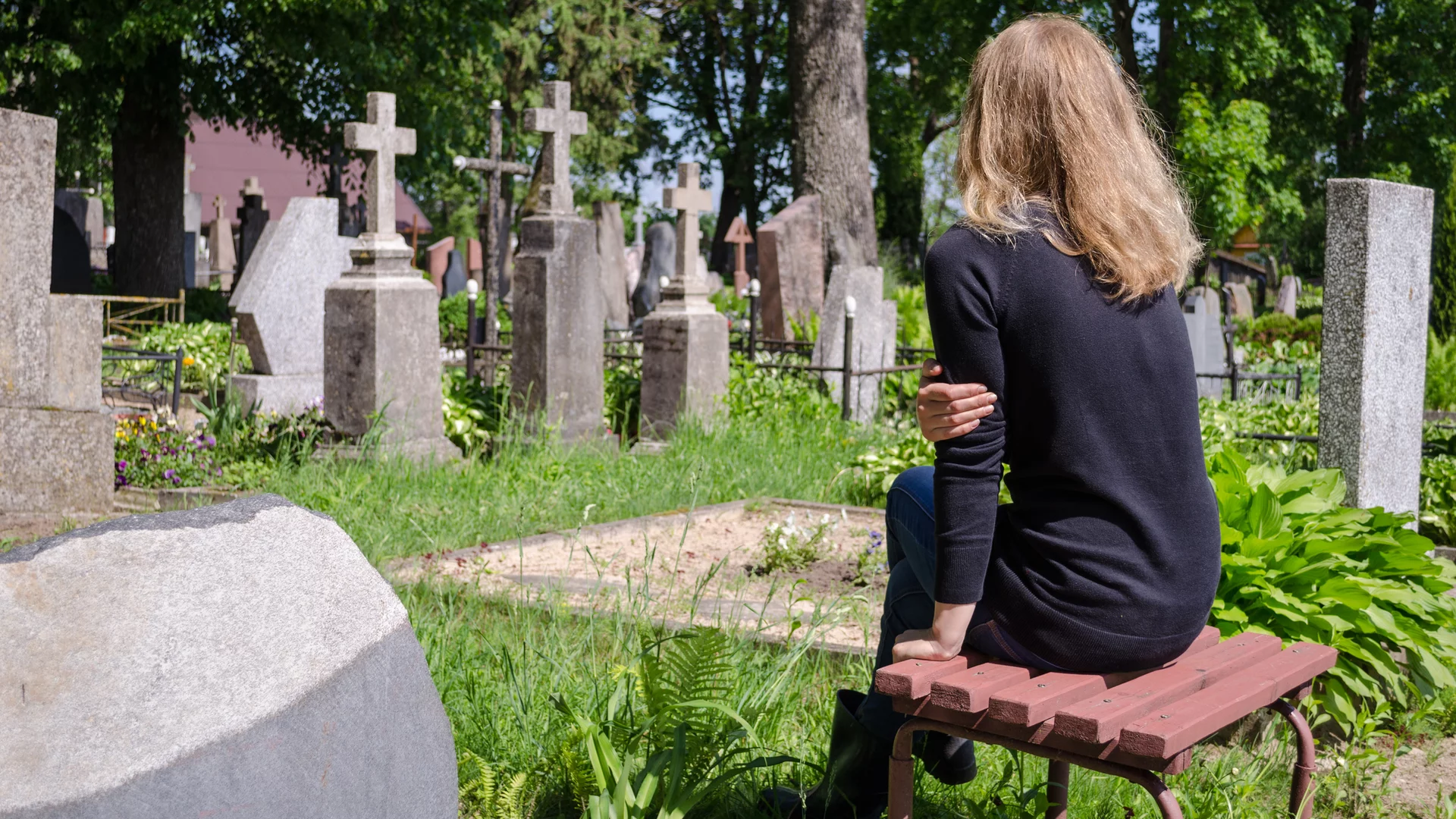 Eine Person mit langen Haaren sitzt auf einer Bank auf einem Friedhof und blickt nachdenklich auf Grabsteine und Kreuze inmitten von grüner Vegetation. Der Tag ist sonnig und es herrscht eine ruhige, friedliche Atmosphäre.