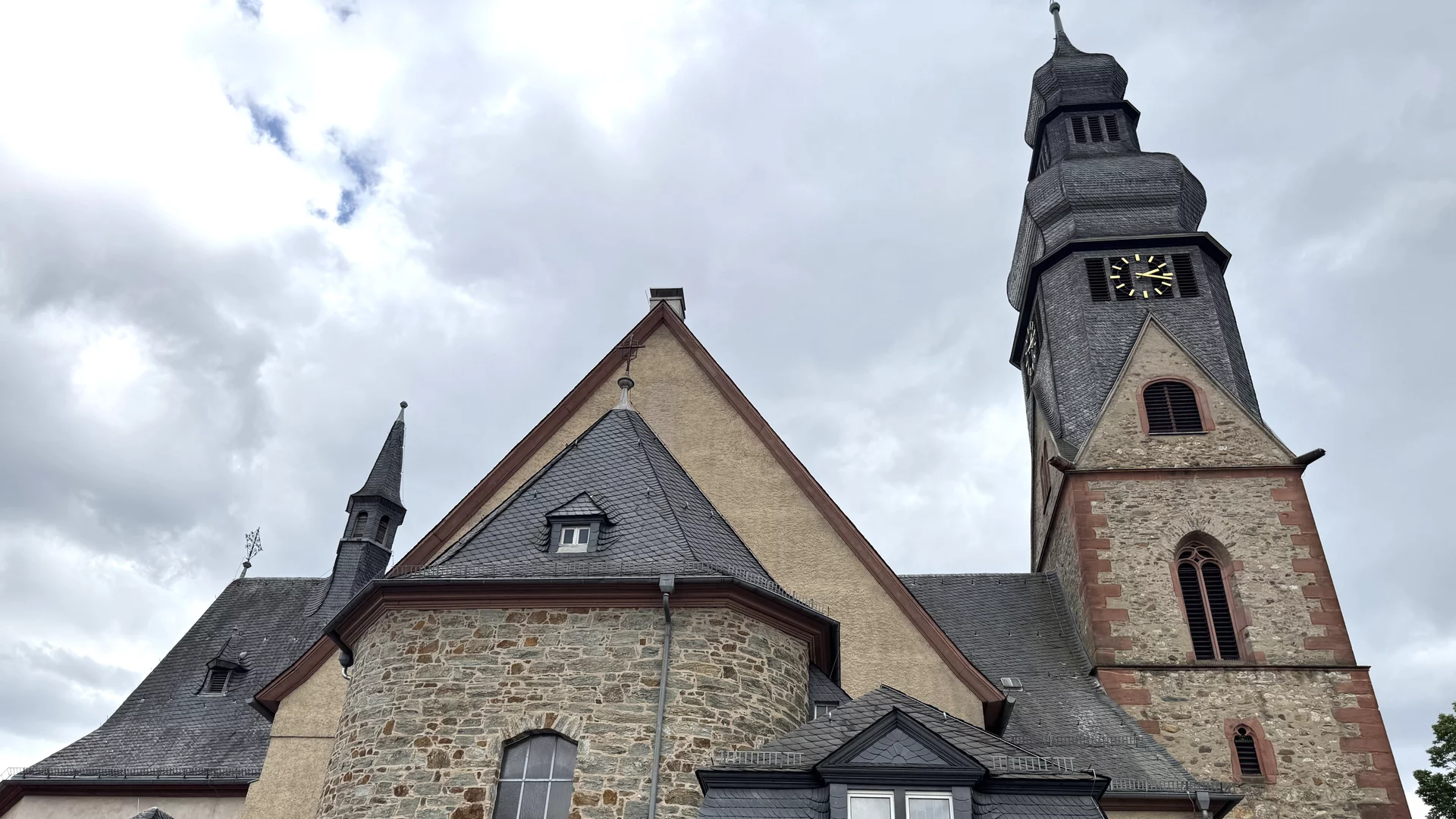Die Abbildung zeigt eine historische Kirche mit einem hohen Glockenturm und verschiedenen Dachformen. Die Fassade besteht aus Stein und ist teilweise mit Holz gestaltet. Der Himmel ist bewölkt, und die Atmosphäre wirkt ruhig und beschaulich.