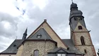 Die Abbildung zeigt eine historische Kirche mit einem hohen Glockenturm und verschiedenen Dachformen. Die Fassade besteht aus Stein und ist teilweise mit Holz gestaltet. Der Himmel ist bewölkt, und die Atmosphäre wirkt ruhig und beschaulich.