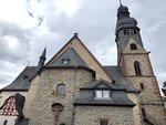 Die Abbildung zeigt eine historische Kirche mit einem hohen Glockenturm und verschiedenen Dachformen. Die Fassade besteht aus Stein und ist teilweise mit Holz gestaltet. Der Himmel ist bewölkt, und die Atmosphäre wirkt ruhig und beschaulich.