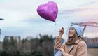 Eine Frau mit langen, lockigen Haaren trägt eine blaue Mütze und einen beigen Mantel. Sie hält einen lila Herzluftballon in der Hand und blickt sanft nach oben. Im Hintergrund sind unscharfe Bäume und eine Brücke zu sehen.