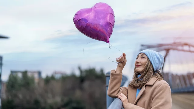 Eine Frau mit langen, lockigen Haaren trägt eine blaue Mütze und einen beigen Mantel. Sie hält einen lila Herzluftballon in der Hand und blickt sanft nach oben. Im Hintergrund sind unscharfe Bäume und eine Brücke zu sehen.