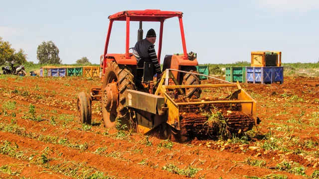 Ein Traktor bearbeitet ein Feld. Eine Person sitzt am Steuer und pflügt den Boden, während im Hintergrund Kisten stehen. Die Erde ist rot, und das Bild vermittelt eine landwirtschaftliche Atmosphäre.