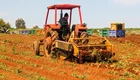 Ein Traktor bearbeitet ein Feld. Eine Person sitzt am Steuer und pflügt den Boden, während im Hintergrund Kisten stehen. Die Erde ist rot, und das Bild vermittelt eine landwirtschaftliche Atmosphäre.