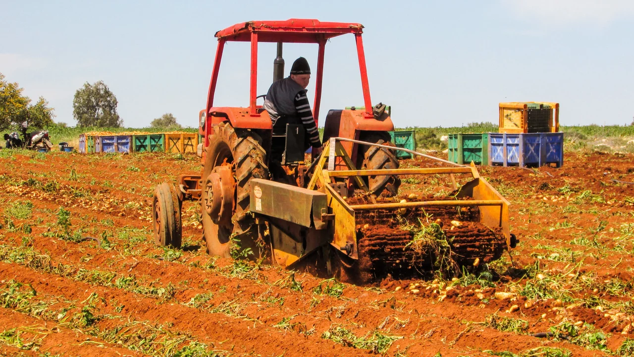 Ein Traktor bearbeitet ein Feld. Eine Person sitzt am Steuer und pflügt den Boden, während im Hintergrund Kisten stehen. Die Erde ist rot, und das Bild vermittelt eine landwirtschaftliche Atmosphäre.