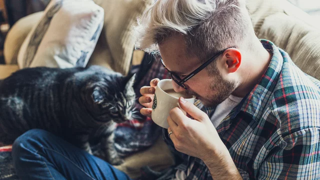 Ein junger Mann mit blonden, kurzen Haaren trinkt aus einer Tasse, während eine neugierige Katze ihn beobachtet. Er sitzt entspannt auf einem Sofa und trägt ein kariertes Hemd. Die Atmosphäre wirkt gemütlich und vertraut.