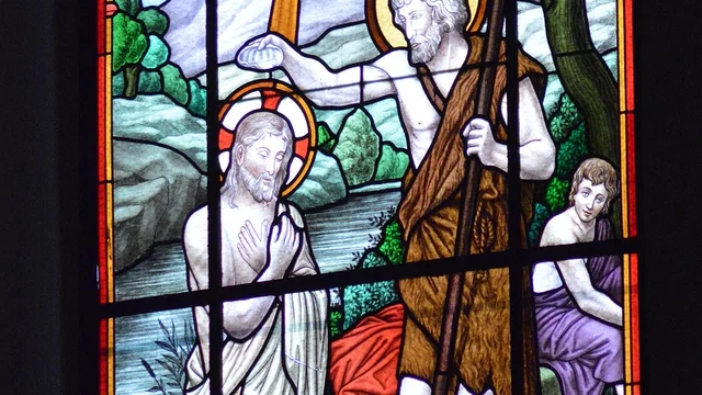 Das Bild zeigt ein buntes Kirchenfenster, in dem Johannes der Täufer Jesus im Wasser tauft. Im Hintergrund sind grüne Hügel und Bäume zu sehen. Eine symbolische Lichtquelle strahlt über der Szene, während eine weitere Figur im Hintergrund aufmerksam zusieht.