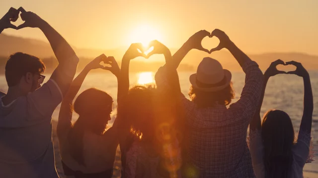 Sechs Personen stehen am Strand bei Sonnenuntergang und formen mit ihren Händen Herzen. Sie blicken auf das Wasser, während die Sonne am Horizont untergeht. Die Atmosphäre vermittelt ein Gefühl von Gemeinschaft und Liebe.