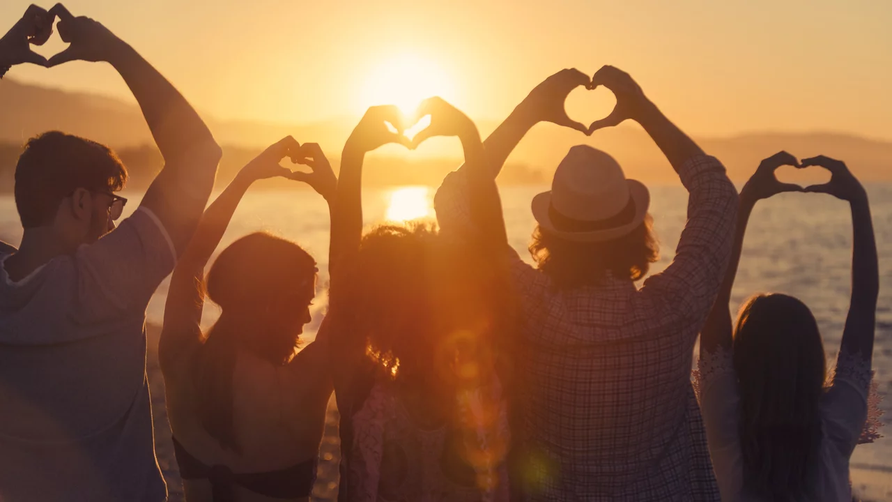 Sechs Personen stehen am Strand bei Sonnenuntergang und formen mit ihren Händen Herzen. Sie blicken auf das Wasser, während die Sonne am Horizont untergeht. Die Atmosphäre vermittelt ein Gefühl von Gemeinschaft und Liebe.