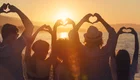 Sechs Personen stehen am Strand bei Sonnenuntergang und formen mit ihren Händen Herzen. Sie blicken auf das Wasser, während die Sonne am Horizont untergeht. Die Atmosphäre vermittelt ein Gefühl von Gemeinschaft und Liebe.
