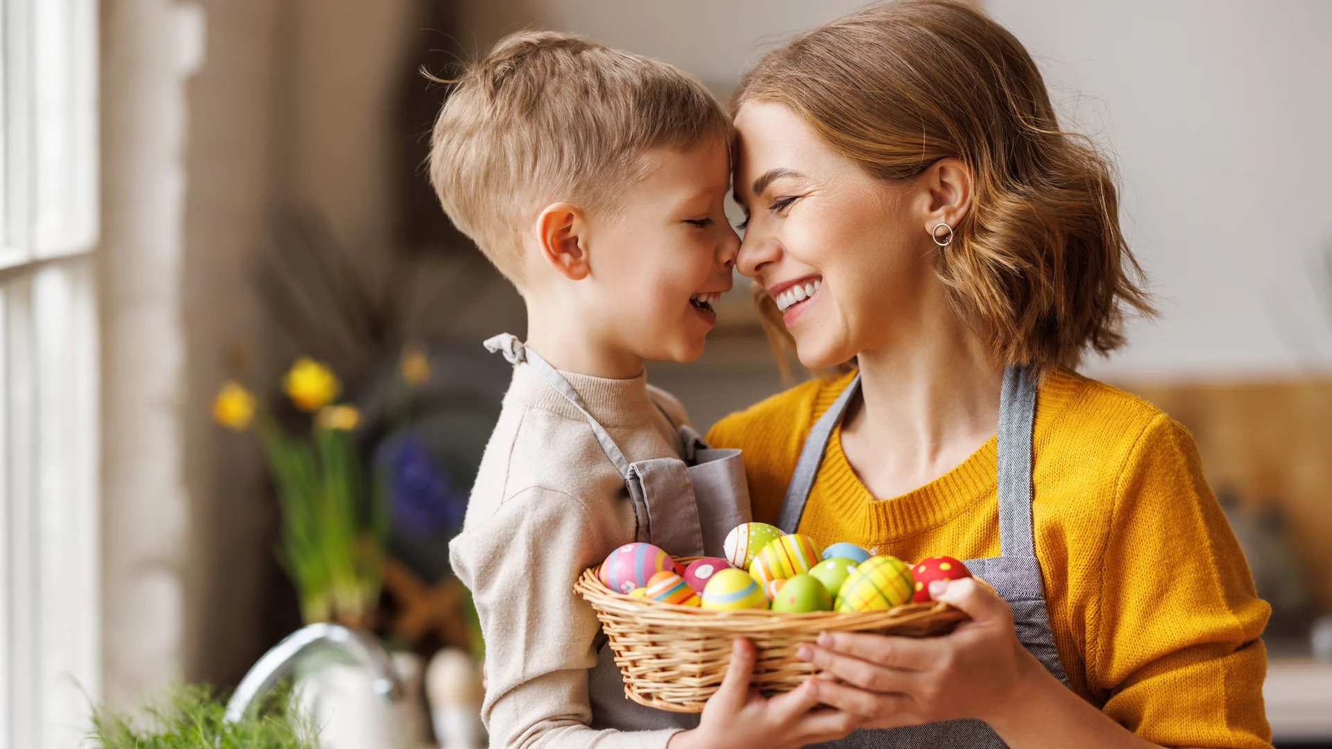 Eine fröhliche Mutter und ihr Sohn lächeln sich an, während sie zusammenstehen. Die Mutter hält einen Korb mit bunten Ostereiern. Die Szene strahlt Wärme und familiäre Freude aus, passend zu den Osterfeierlichkeiten.