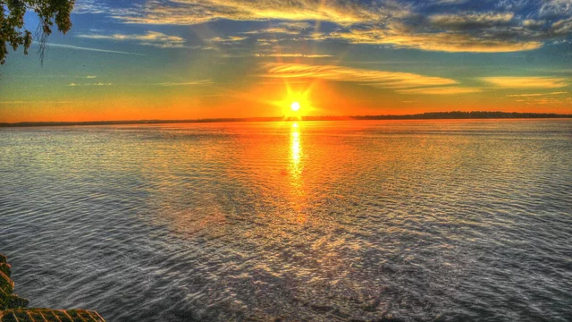 Ein Sonnenuntergang über einem ruhigen Gewässer, das in warmen Farbtönen von Orange und Gelb leuchtet. Die Wolken am Himmel reflektieren das Licht, während der Sonnenstrahl sanft auf der Wasseroberfläche glitzert. Ein friedliches und malerisches Naturszenario.