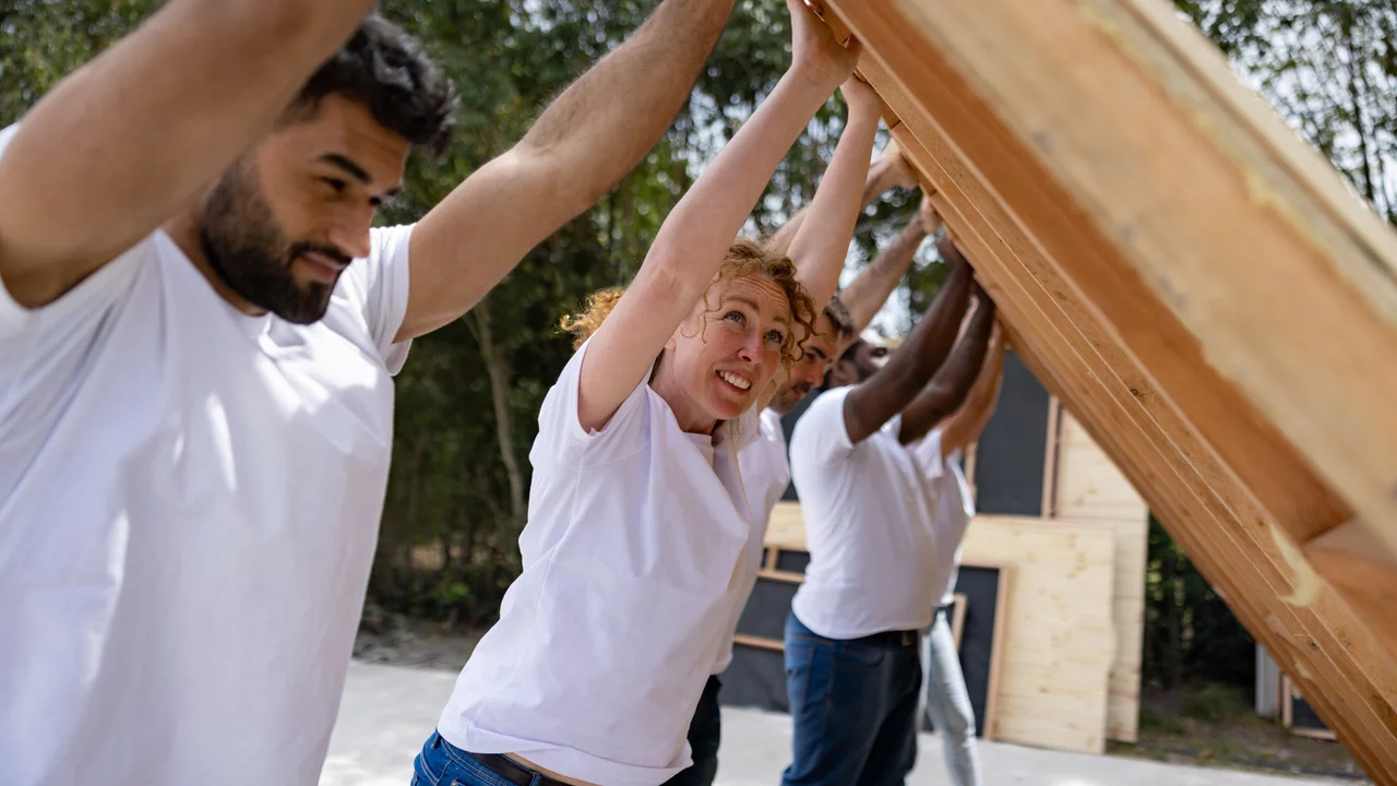 Eine Gruppe von vier Personen arbeitet gemeinsam daran, ein Dach zu heben. Sie tragen weiße T-Shirts und zeigen Konzentration und Teamarbeit. Die Szene vermittelt ein Gefühl der Zusammenarbeit und des handwerklichen Schaffens in einer ländlichen Umgebung.