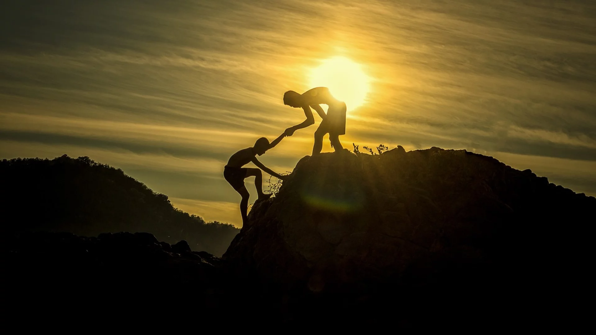 Zwei Silhouetten von Personen stehen auf einem Felsen und helfen sich gegenseitig nach oben. Im Hintergrund ist die Sonne zu sehen, die am Horizont aufgeht. Die Szene vermittelt ein Gefühl von Zusammenarbeit und Unterstützung.