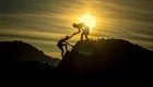 Zwei Silhouetten von Personen stehen auf einem Felsen und helfen sich gegenseitig nach oben. Im Hintergrund ist die Sonne zu sehen, die am Horizont aufgeht. Die Szene vermittelt ein Gefühl von Zusammenarbeit und Unterstützung.