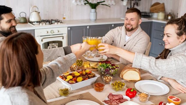 Eine Gruppe von vier Personen sitzt an einem Tisch und stößt mit Getränken an. Auf dem Tisch sind verschiedene Speisen wie Obst, Gemüse, Aufschnitt und Brot angerichtet. Die Atmosphäre ist freundlich und gesellig.