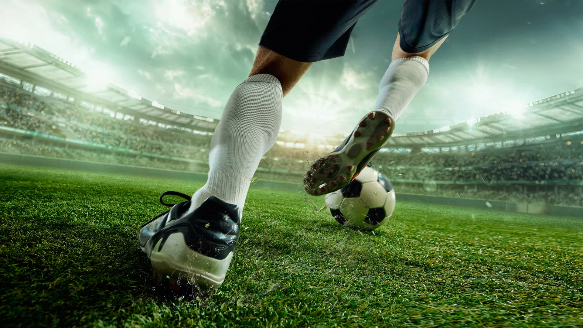 Ein Fußballspieler tritt den Ball auf einem grünen Spielfeld. Die Szene zeigt die Beine des Spielers und den Ball, während das Stadion im Hintergrund sichtbar ist. Das Licht ist dramatisch, was eine spannende Stimmung vermittelt.
