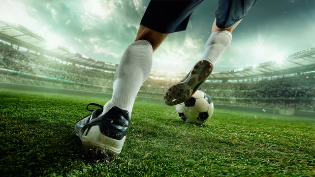 Ein Fußballspieler tritt den Ball auf einem grünen Spielfeld. Die Szene zeigt die Beine des Spielers und den Ball, während das Stadion im Hintergrund sichtbar ist. Das Licht ist dramatisch, was eine spannende Stimmung vermittelt.