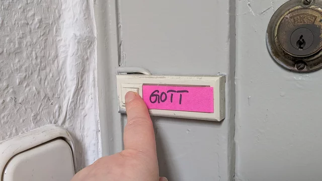 Eine Hand drückt auf einen Klingelknopf an einer Tür, der mit einem pinken Aufkleber beschriftet ist. Auf dem Aufkleber steht das Wort "GOTT". Im Hintergrund ist ein Türschloss sichtbar.