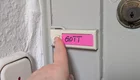 Eine Hand drückt auf einen Klingelknopf an einer Tür, der mit einem pinken Aufkleber beschriftet ist. Auf dem Aufkleber steht das Wort "GOTT". Im Hintergrund ist ein Türschloss sichtbar.