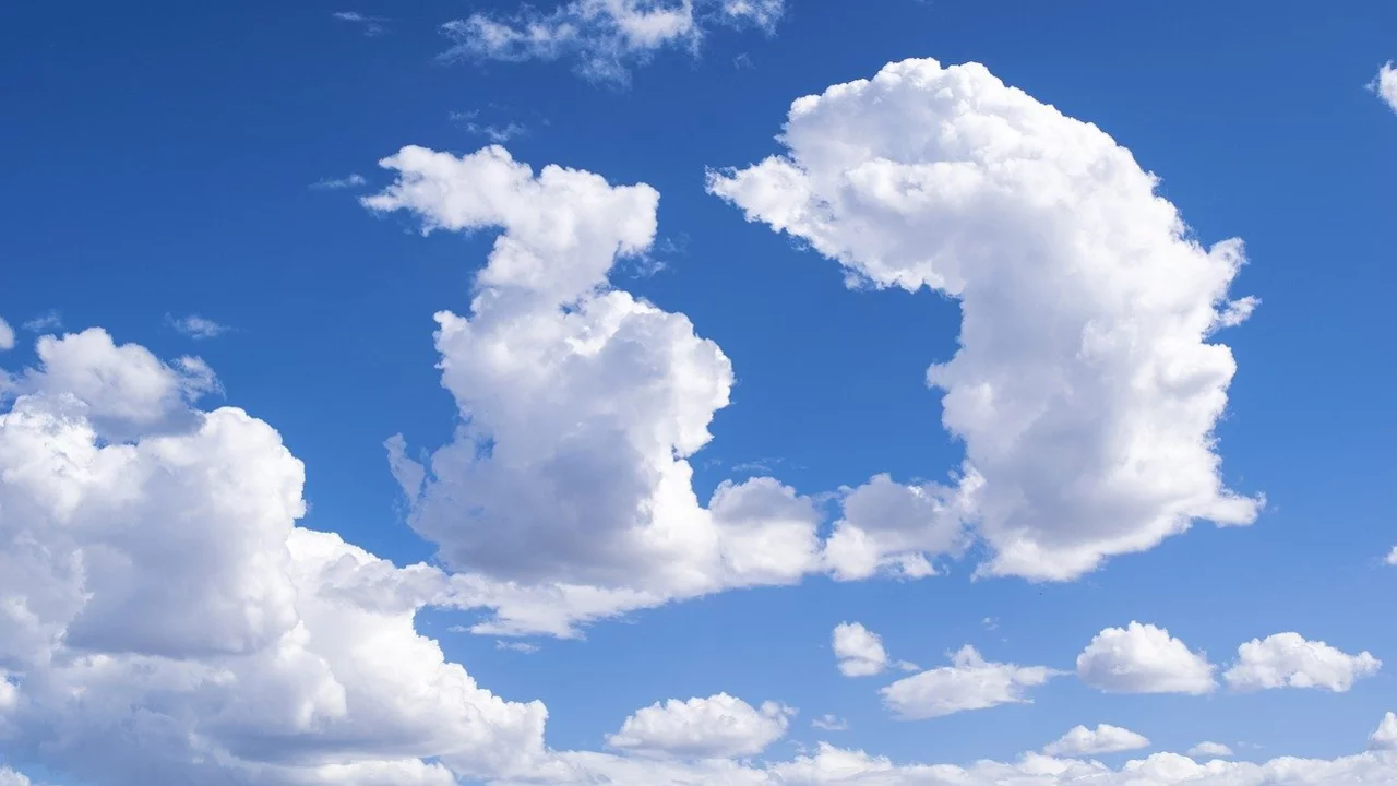 Der Himmel ist klar und blau, durchzogen von verschiedenen Wolkenformen. Einige Wolken bilden markante, geflügelte Gestalten, die an Tiere oder Menschen erinnern. Die insgesamt helle und freundliche Atmosphäre strahlt Ruhe aus.