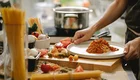 Ein Koch richtet einen Teller mit Spaghetti und Tomatensauce an. Neben dem Teller befinden sich frische Zutaten wie Tomaten, Gemüse und verschiedene Pasta-Sorten. Im Hintergrund steht ein Topf auf dem Herd.