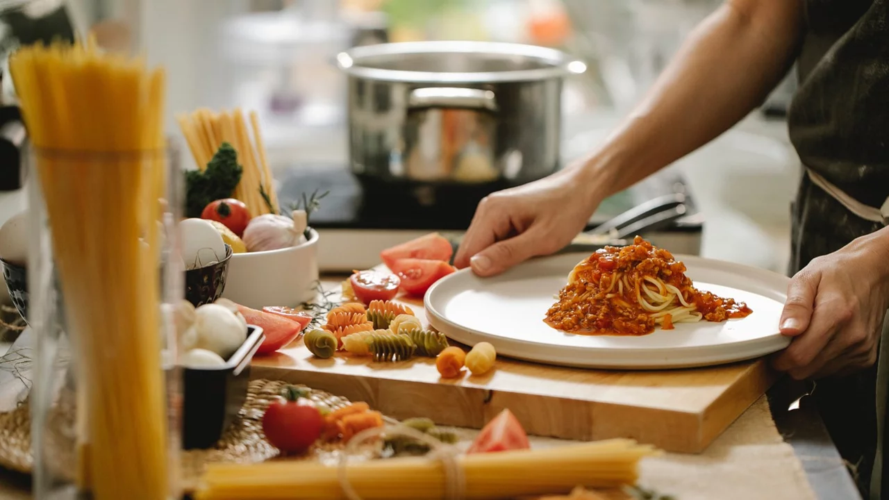 Ein Koch richtet einen Teller mit Spaghetti und Tomatensauce an. Neben dem Teller befinden sich frische Zutaten wie Tomaten, Gemüse und verschiedene Pasta-Sorten. Im Hintergrund steht ein Topf auf dem Herd.