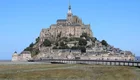 Eine beeindruckende mittelalterliche Abtei steht auf einem Hügel, umgeben von Wasser und einer weiten Landschaft. Die Architektur ist detailliert, mit Türmen und Mauern, die die Geschichte widerspiegeln. Ein Weg führt zu der Abtei, die bei Flut teilweise von Wasser umgeben ist.