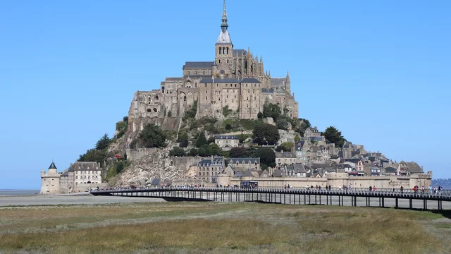 Eine beeindruckende mittelalterliche Abtei steht auf einem Hügel, umgeben von Wasser und einer weiten Landschaft. Die Architektur ist detailliert, mit Türmen und Mauern, die die Geschichte widerspiegeln. Ein Weg führt zu der Abtei, die bei Flut teilweise von Wasser umgeben ist.