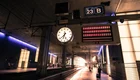 Ein Bahnhof mit einem Taktik-Display und einer großen Uhr. Im Hintergrund sind Reisende zu sehen, die an Gleis 23 warten. Die Beleuchtung ist gedimmt, und die Wände sind mit modernen Materialien gestaltet. Das Bild vermittelt eine ruhige, wartende Atmosphäre.