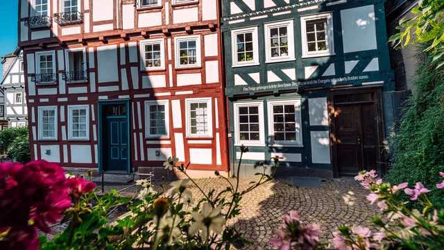 Die Aufnahme zeigt ein Straßenpanorama mit Fachwerkhäusern in verschiedenen Farbtönen, einschließlich rot und blau. Umgeben von Blumen und grünem Laub verleihen sie der Szenerie einen charmanten, historischen Charakter. Ein sonniger Tag sorgt für eine freundliche Atmosphäre.