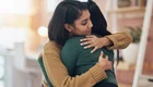 Zwei Frauen umarmen sich herzlich in einem freundlichen Raum. Eine trägt einen beigen Pullover, die andere ein grünes Oberteil. Die Umarmung vermittelt Nähe und Unterstützung in einem momentanen emotionalen Austausch.