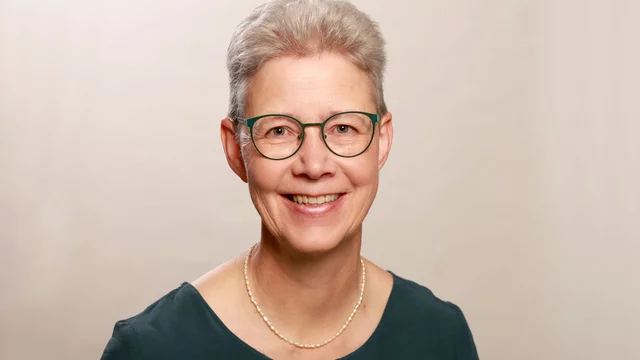 Porträt einer lächelnden Frau mit grauem, kurzem Haar und einer modischen Brille. Sie trägt ein dunkles Oberteil und eine Perlenkette. Der Hintergrund ist weich und neutral gehalten.