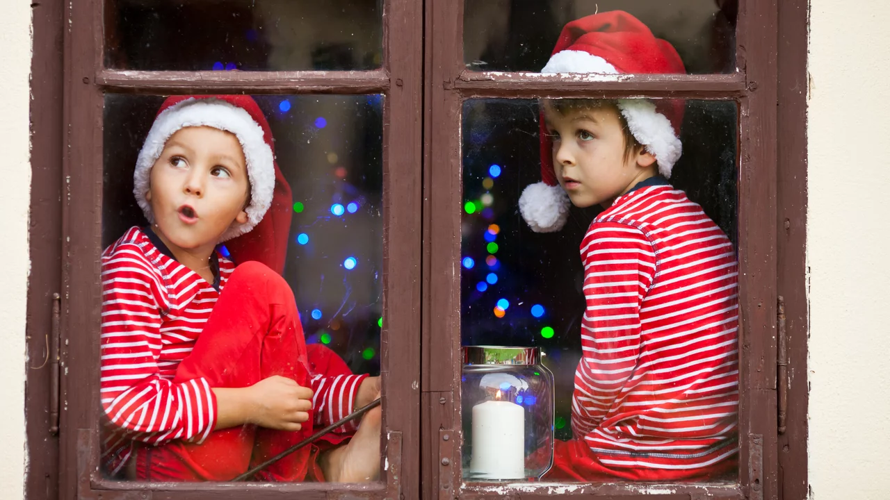 An einem Fenster sitzen zwei Kinder auf dem Fensterbrett. Sie haben rot-weiße Schlafanzüge an und rote Nikolausmützen auf und schauen erwartungsvoll nach draußen. 
