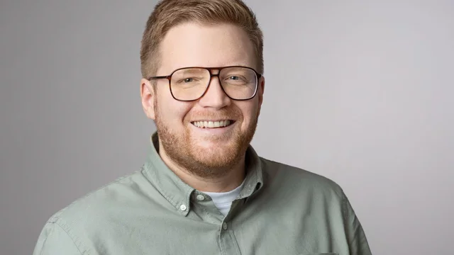 Ein lächelnder Mann mit mittellangen, blonden Haaren und einer Brille steht in einem hellen, neutralen Hintergrund. Er trägt ein graugrünliches Hemd und sieht freundlich aus.