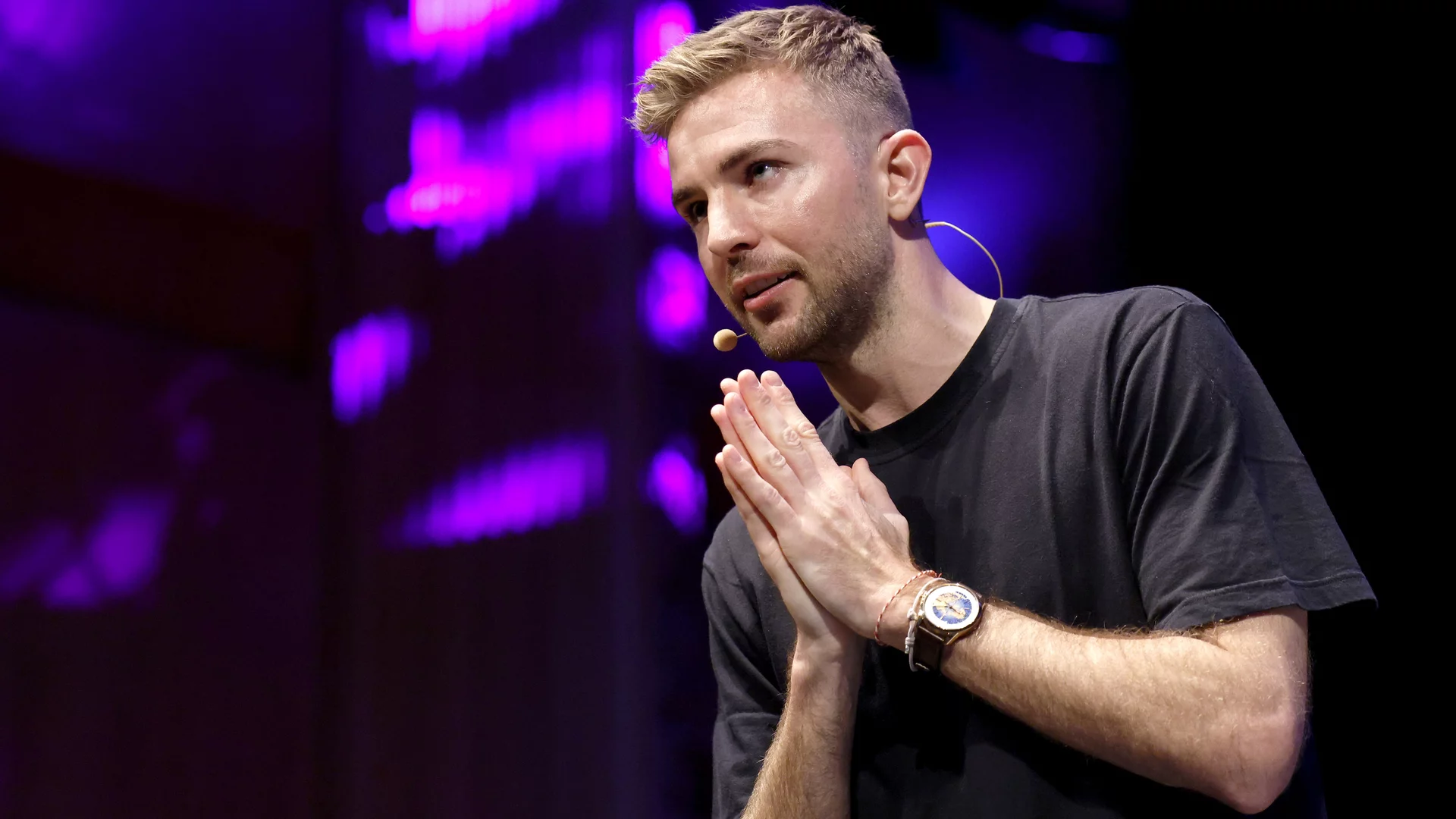 Ein Mann in einem schwarzen T-Shirt steht auf einer Bühne, hält seine Hände in einer betenden Geste. Er spricht wahrscheinlich vor einem Publikum, während bunte Lichter im Hintergrund leuchten.