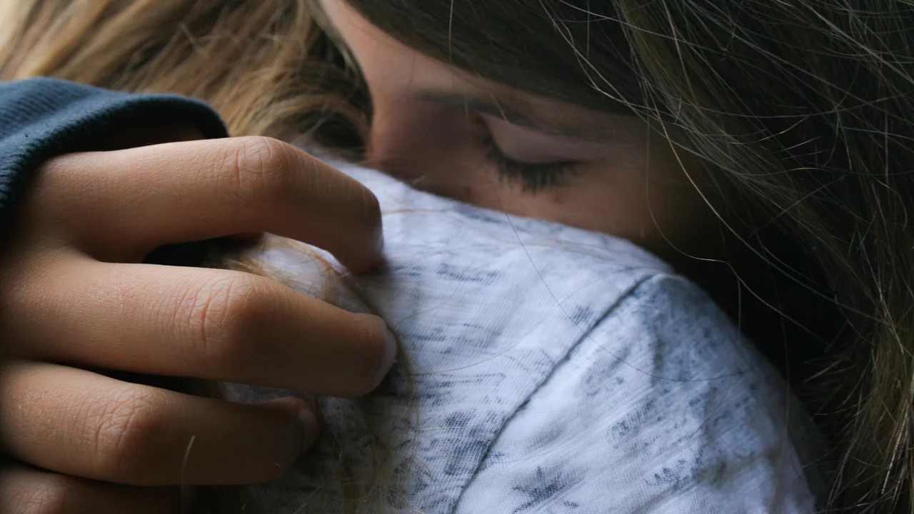 Zwei Personen umarmen sich eng. Eine der Personen hat den Kopf an der Schulter der anderen gelehnt und zeigt eine friedliche, beruhigte Miene. Die Umarmung vermittelt Gefühle von Nähe und Trost.