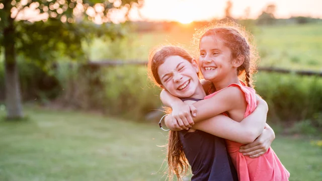 Zwei Mädchen umarmen sich lächelnd im Freien, während die Sonne im Hintergrund untergeht. Sie stehen auf einer grünen Wiese und strahlen Freude und Freundschaft aus.