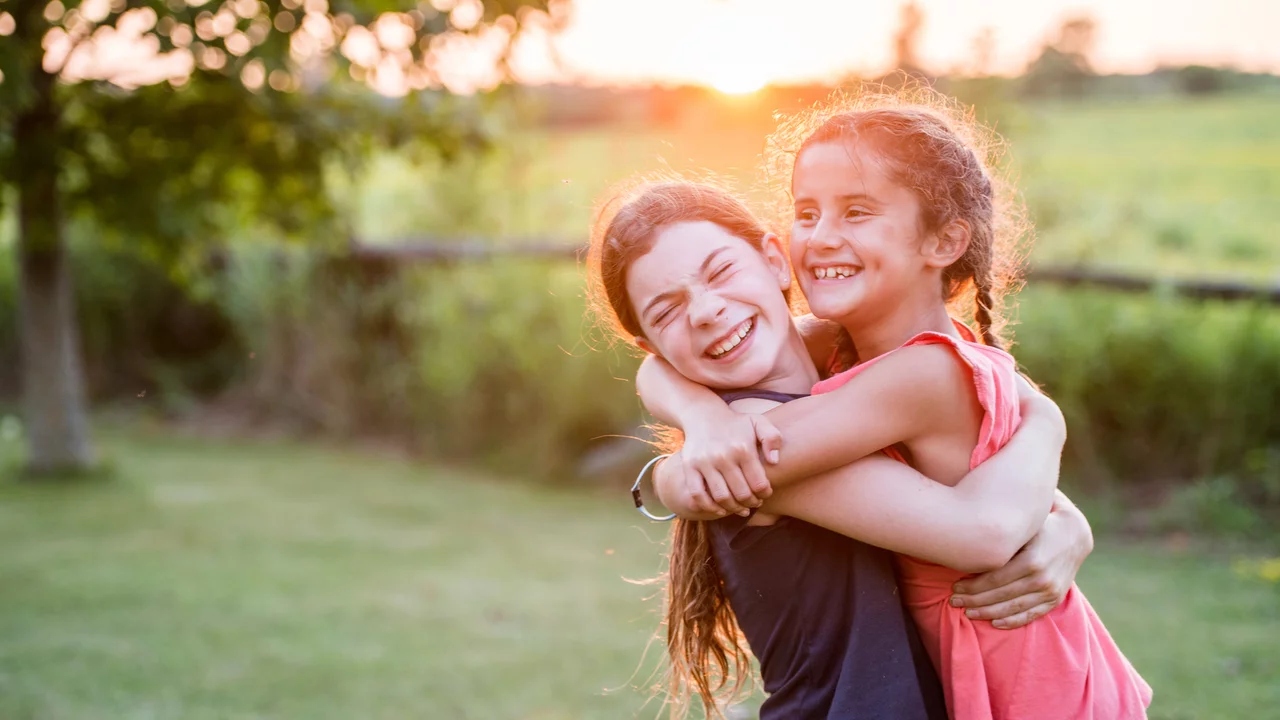 Zwei Mädchen umarmen sich lächelnd im Freien, während die Sonne im Hintergrund untergeht. Sie stehen auf einer grünen Wiese und strahlen Freude und Freundschaft aus.